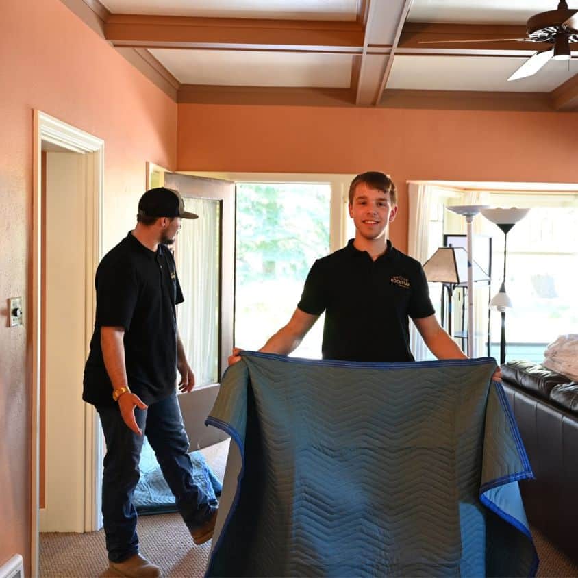 Two movers from Rockstar Moving Company packing a large item in a living room.